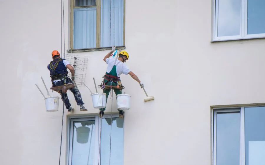 Two exterior painters on harnesses rolling fresh paint onto a cream stucco wall, West Palm Beach, FL