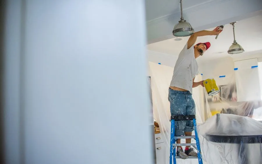Painter on a ladder cutting in a ceiling line during an interior repaint in West Palm Beach, FL, furniture masked with plastic