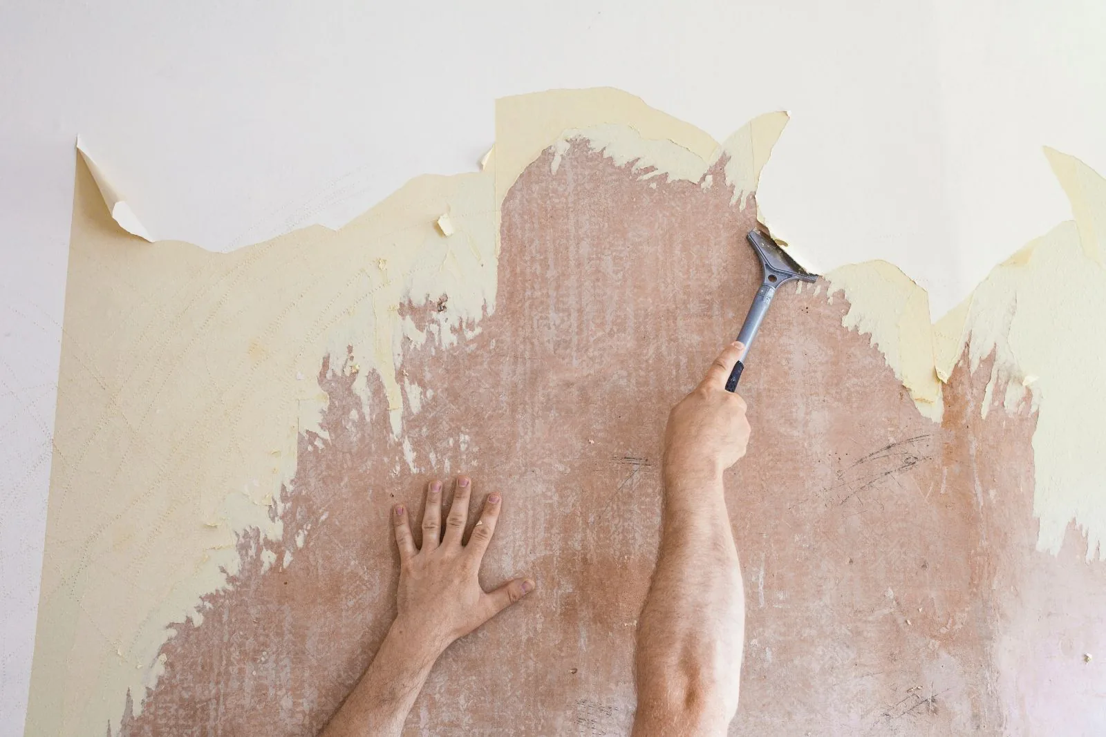 Scraping old yellowed wallpaper off a wall with a long-handled scraper, bare wall showing underneath, West Palm Beach, FL